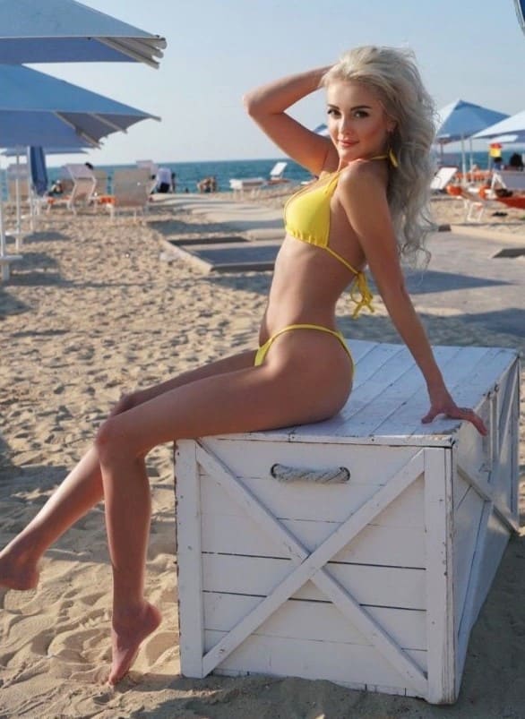 A blonde woman in a yellow two-piece bikini sits on a white wooden crate on the sand, one hand behind her head, with the sea in the background.