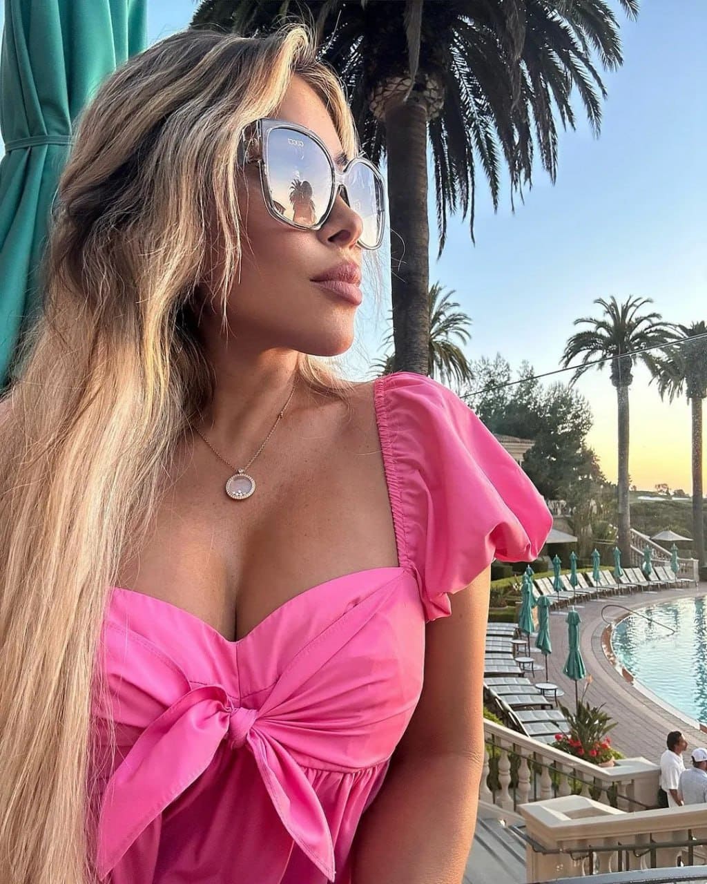 A woman in a bright pink bow top and reflective sunglasses stands on a balcony overlooking a pool area with palm trees.