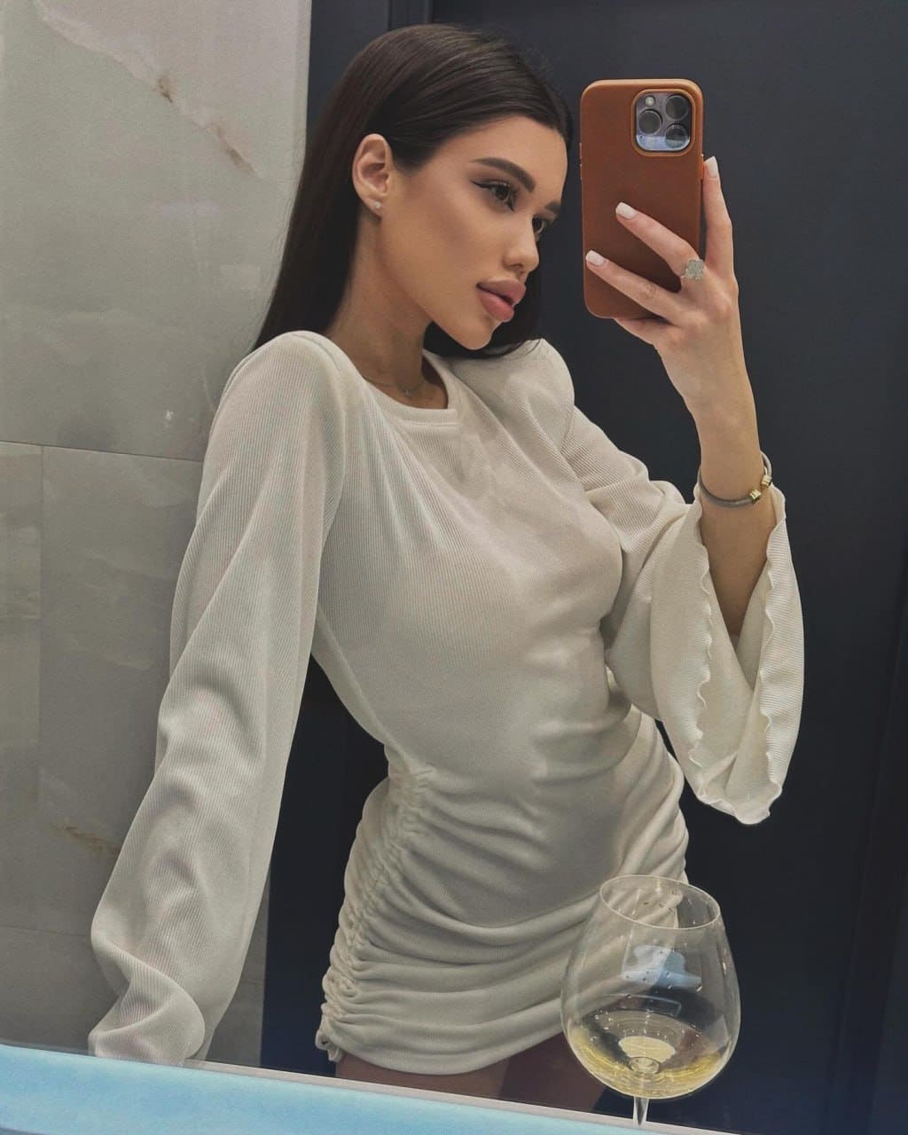 A mirror selfie shows a woman in a fitted white dress with ruffled cuffs, holding a phone and a glass of white wine on a marble counter.