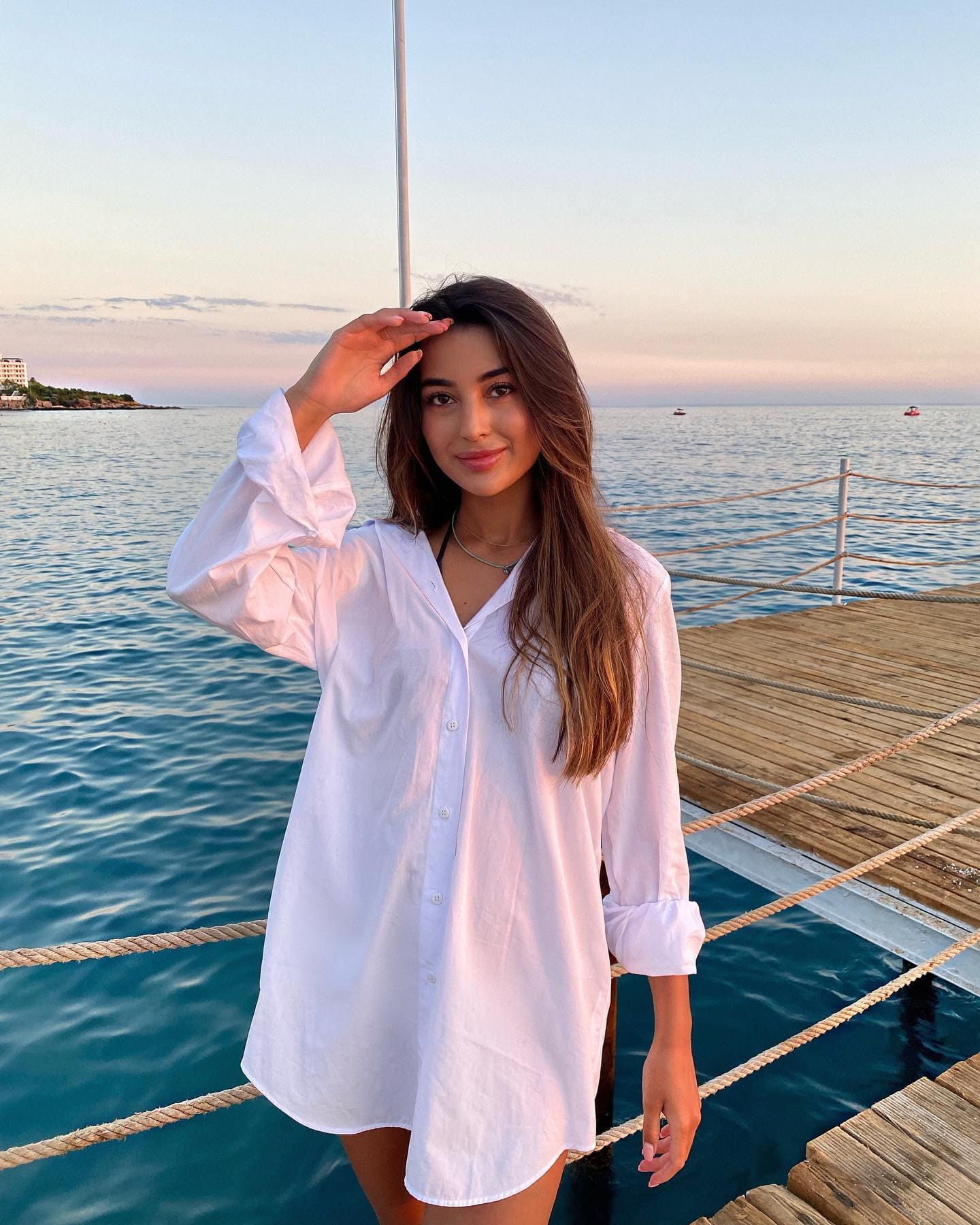 A woman stands on a wooden dock by the sea, wearing an oversized white button-up shirt with rolled sleeves and her hand raised to her forehead.
