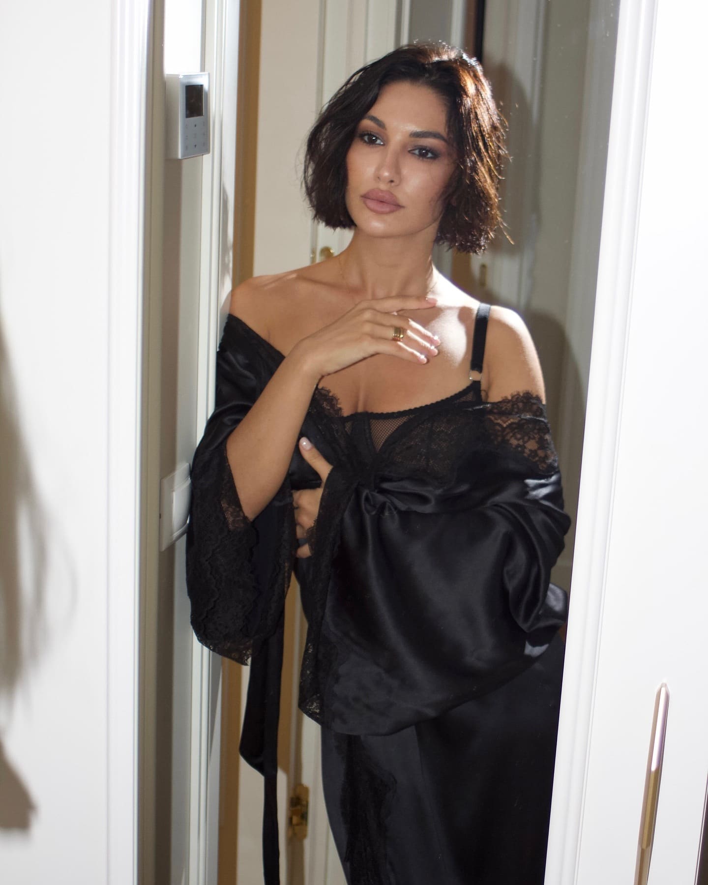 Sofia stands in a doorway wearing a black lace and satin robe over a black lace bralette, with one hand raised near her chest.