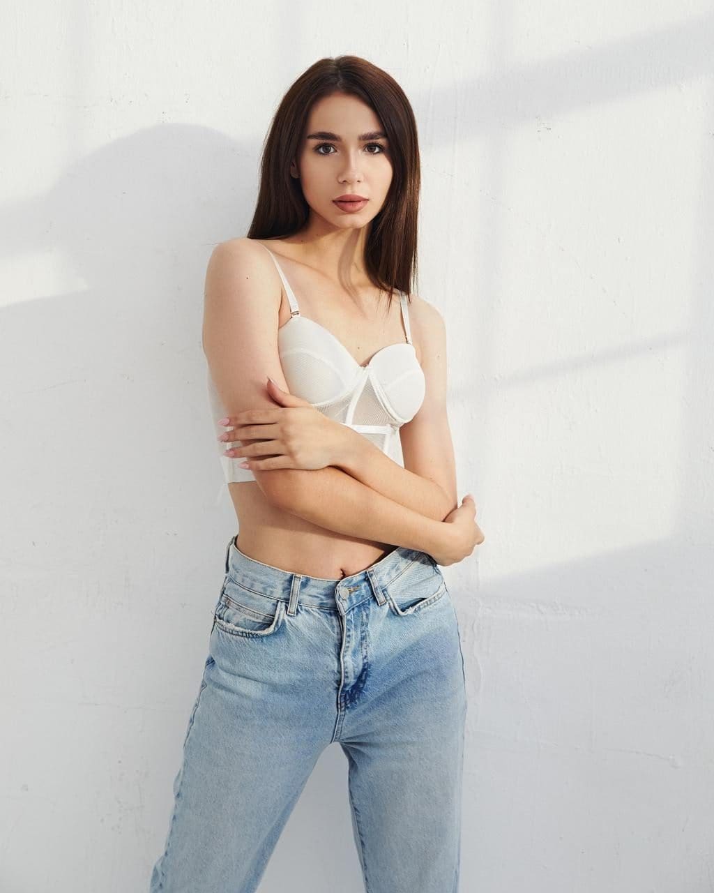 Milly stands against a white wall in a white bra and light blue jeans, arms crossed over her torso.
