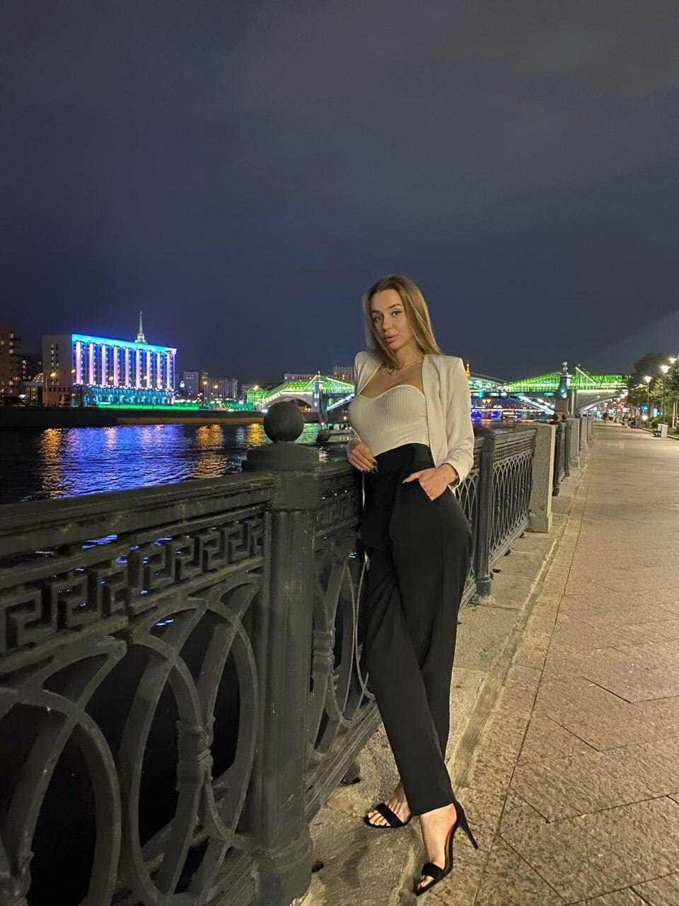 Woman in a white cropped blazer and black trousers leans on a railing by a river at night, city lights behind.