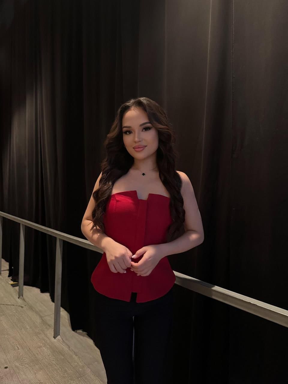 Standing before a black curtain, she wears a red strapless peplum top and black pants, with her hands clasped in front of her waist.