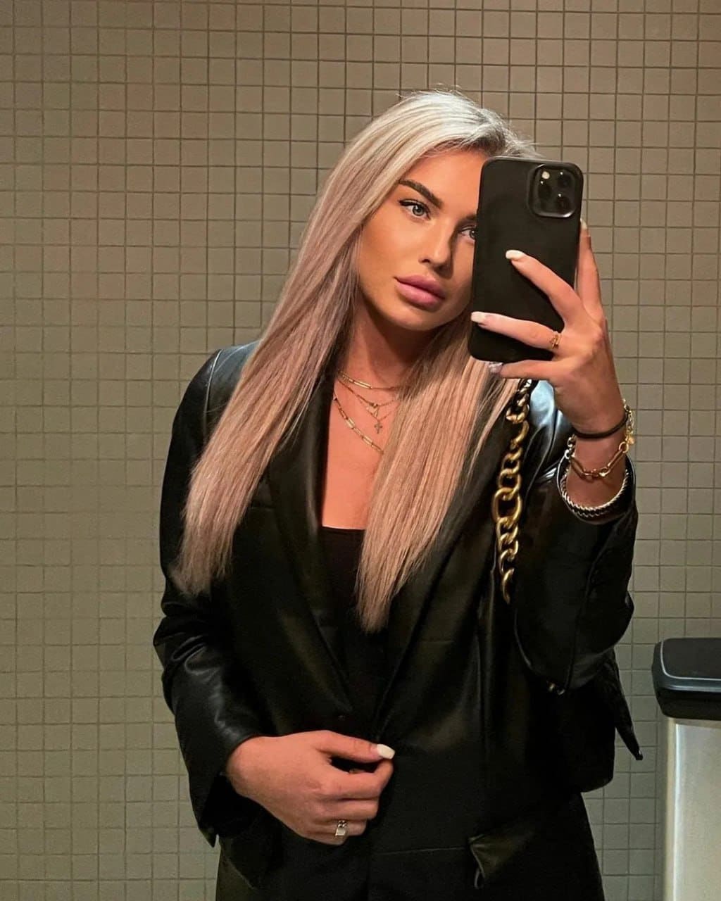 Mirror selfie of a platinum-blonde woman in a black leather blazer and layered jewelry, standing before a beige tiled wall.