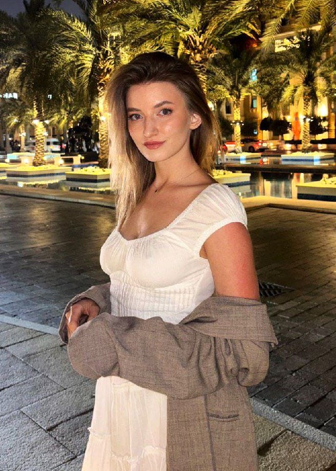 A woman with long blonde hair stands outdoors at night near palm trees and a fountain, wearing a white low-cut top and grey blazer.