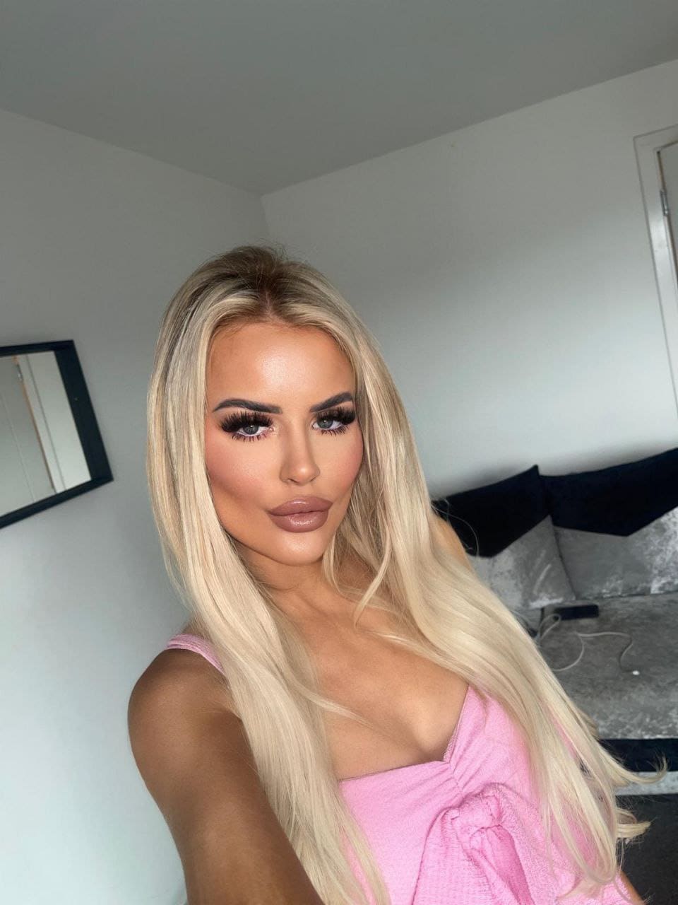 A woman with straight blonde hair wears a pink sleeveless top indoors, standing near a white wall and dark sofa.