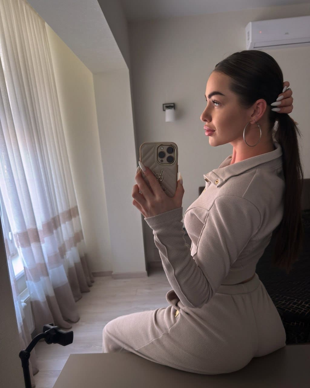 Sitting in profile on a table in a beige outfit, holding a phone and touching her hair near sheer curtains.