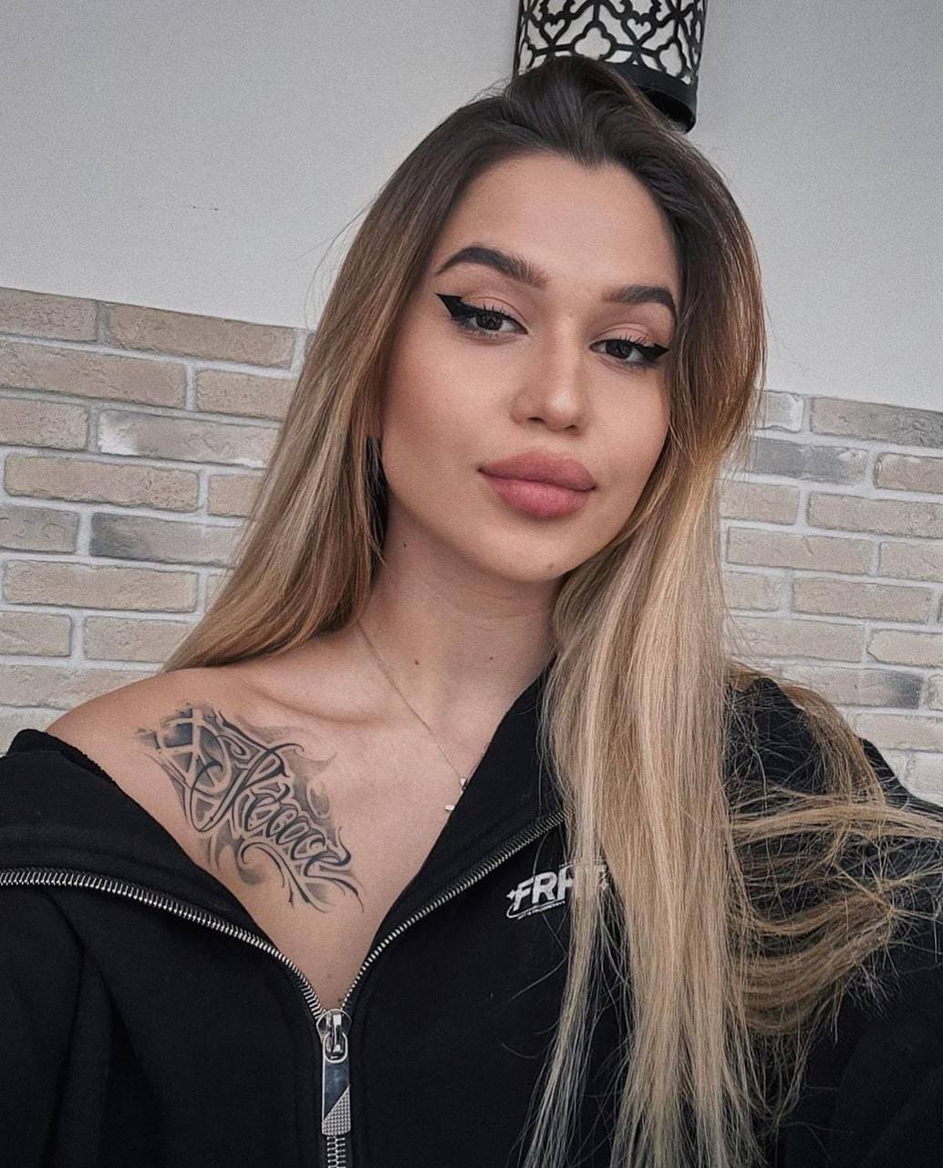Mia stands in a black zip-up hoodie against a light brick wall, with long blonde hair and a shoulder tattoo visible.