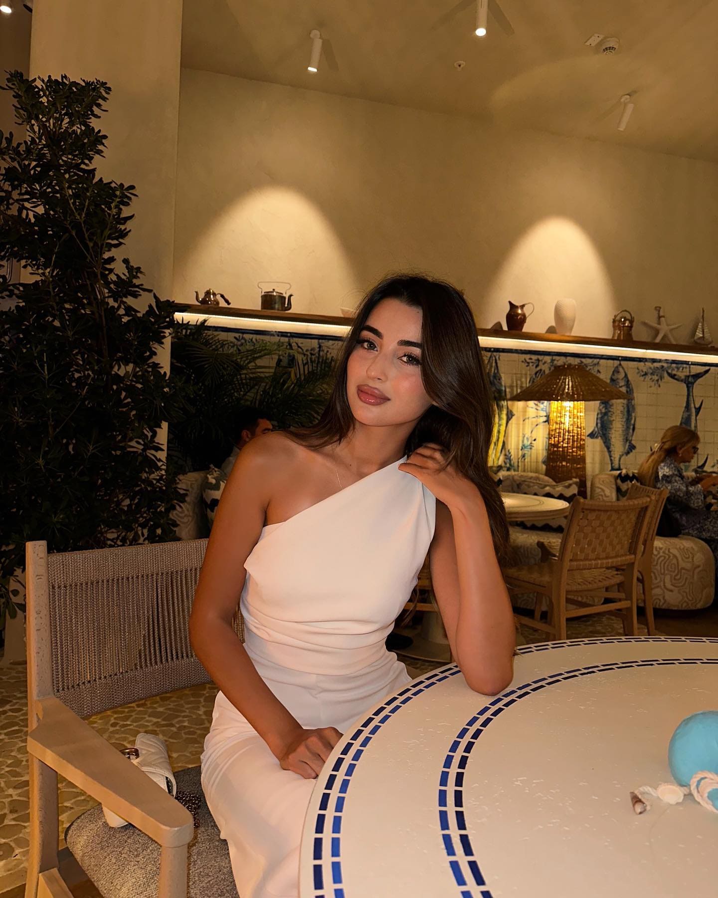 A woman in a white one-shoulder dress sits at a round tiled table in a restaurant, resting one hand on her shoulder.