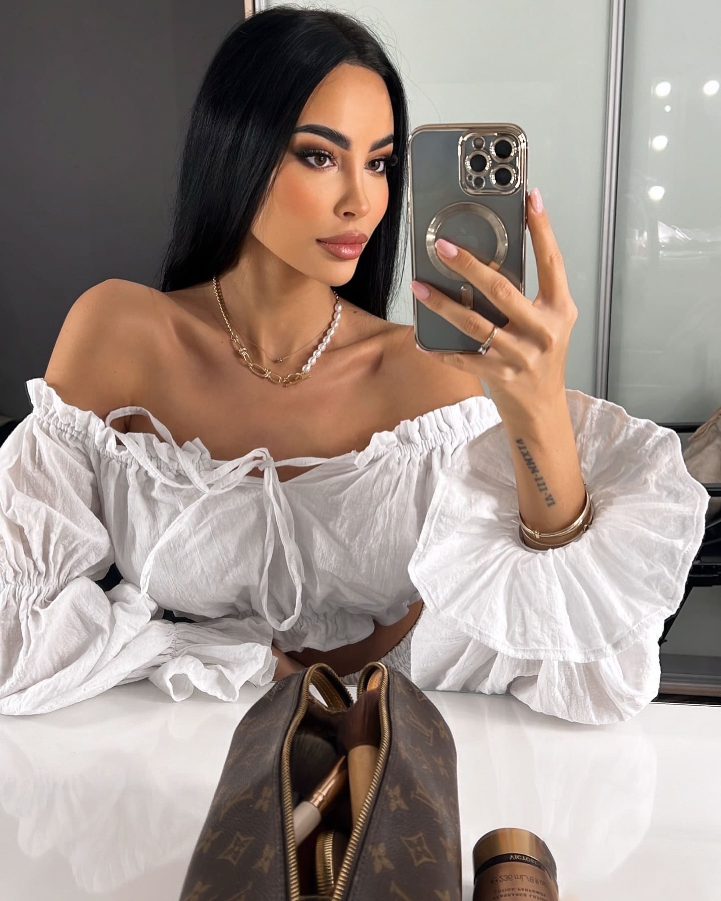 In a mirror selfie, she wears a white ruffled off-shoulder blouse and layered necklaces, with a handbag nearby.