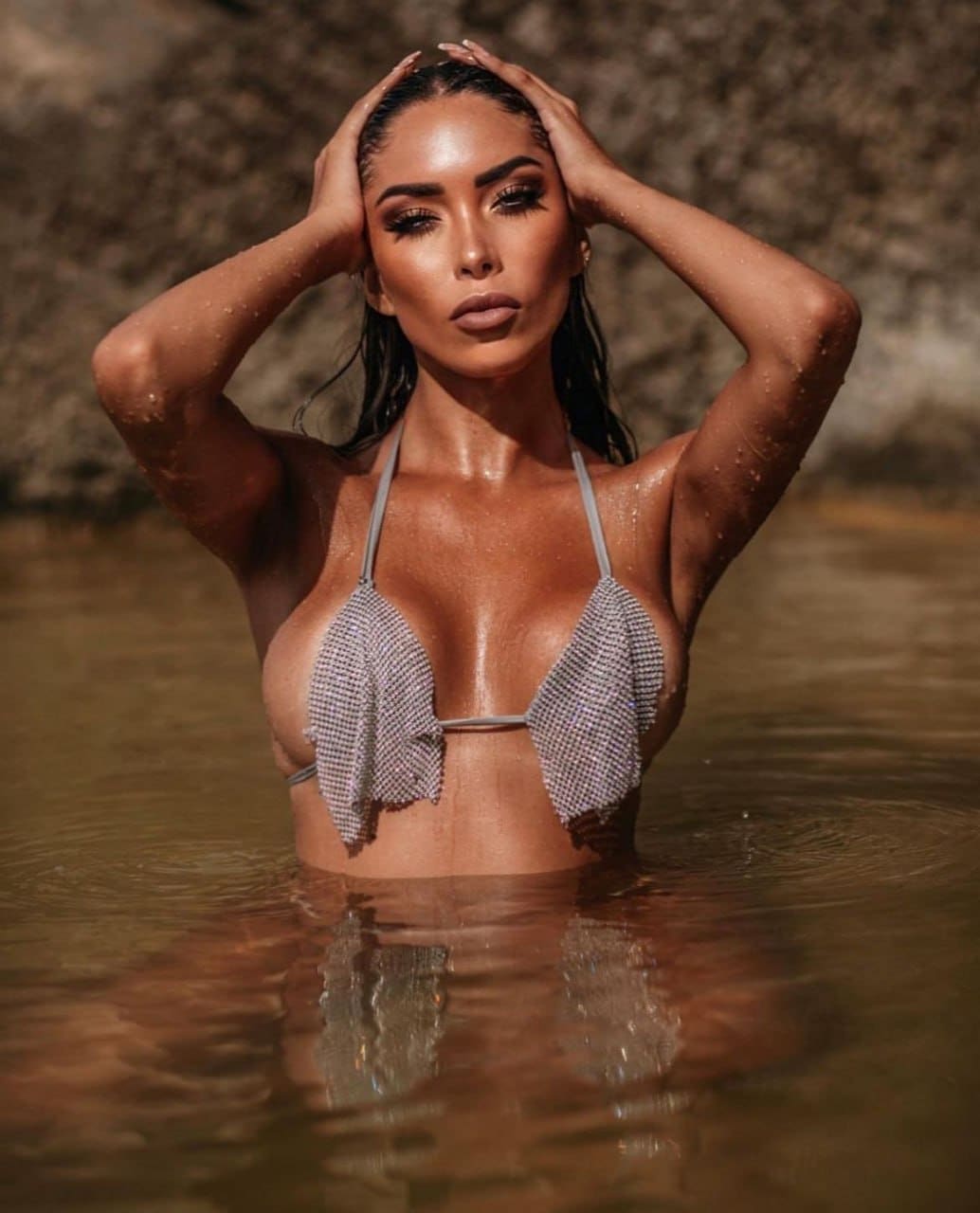 A woman with wet hair stands in shallow water wearing a textured light bikini top, hands on her head, rocky background.