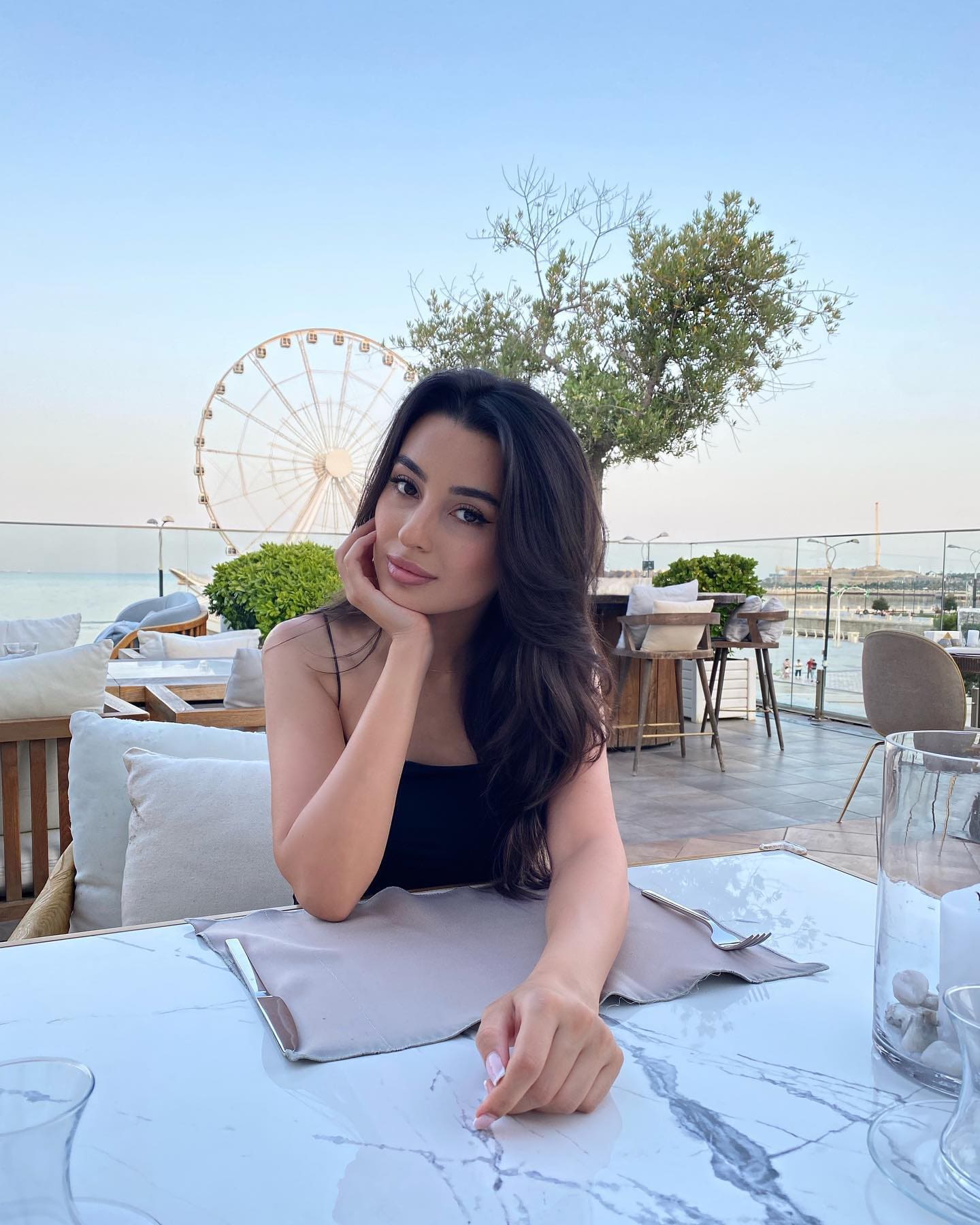 Kantira sits at a marble table on a terrace, chin on hand, wearing a black sleeveless top with a Ferris wheel and sea behind her.