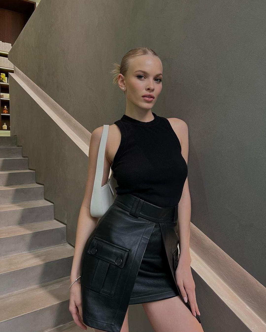 A blonde woman stands on concrete stairs in a black sleeveless top and leather skirt with a wide belt, carrying a white shoulder bag.