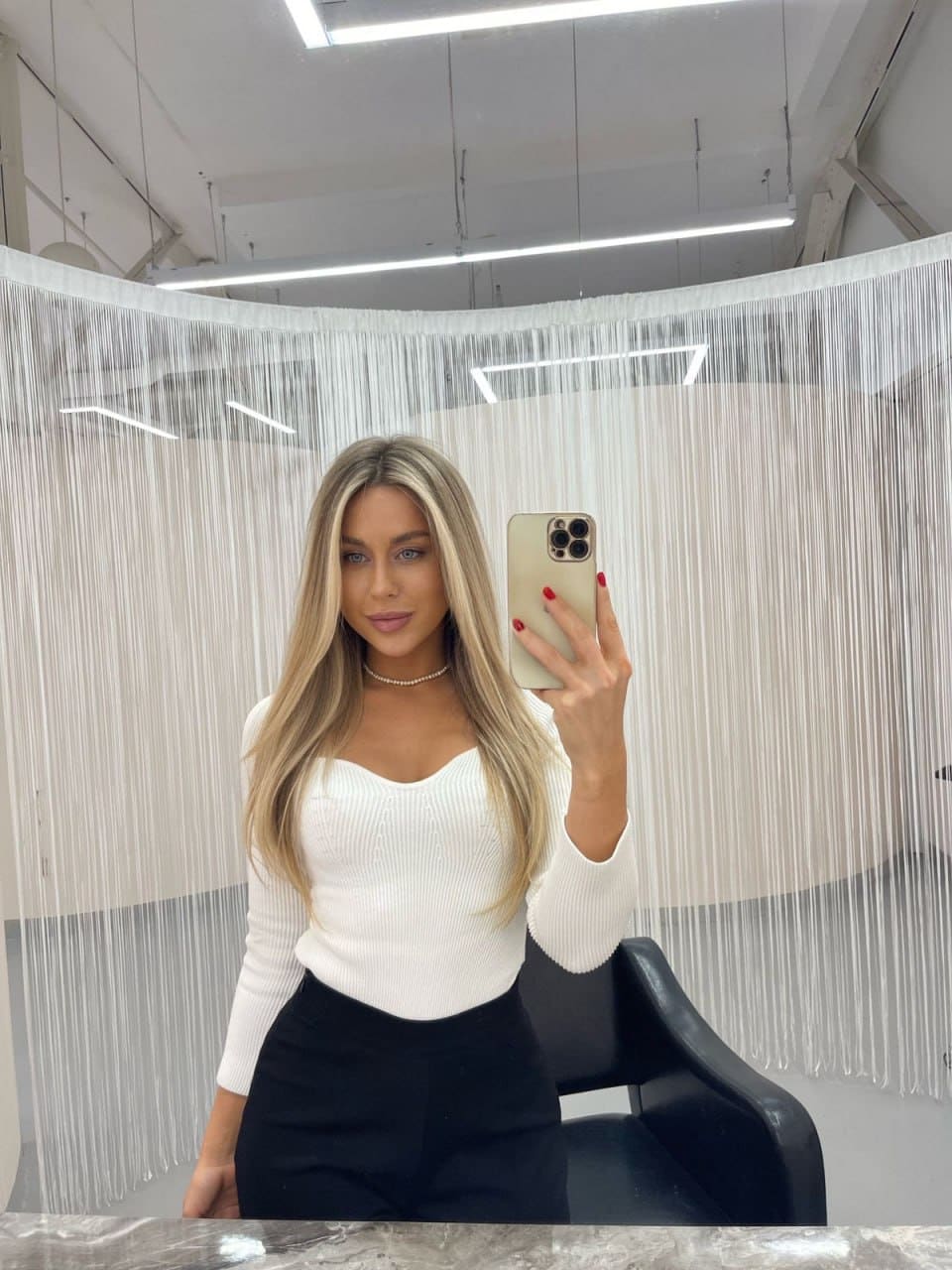 Lida takes a mirror selfie in a white ribbed long-sleeve top and black skirt, standing before a white beaded curtain in a bright room.