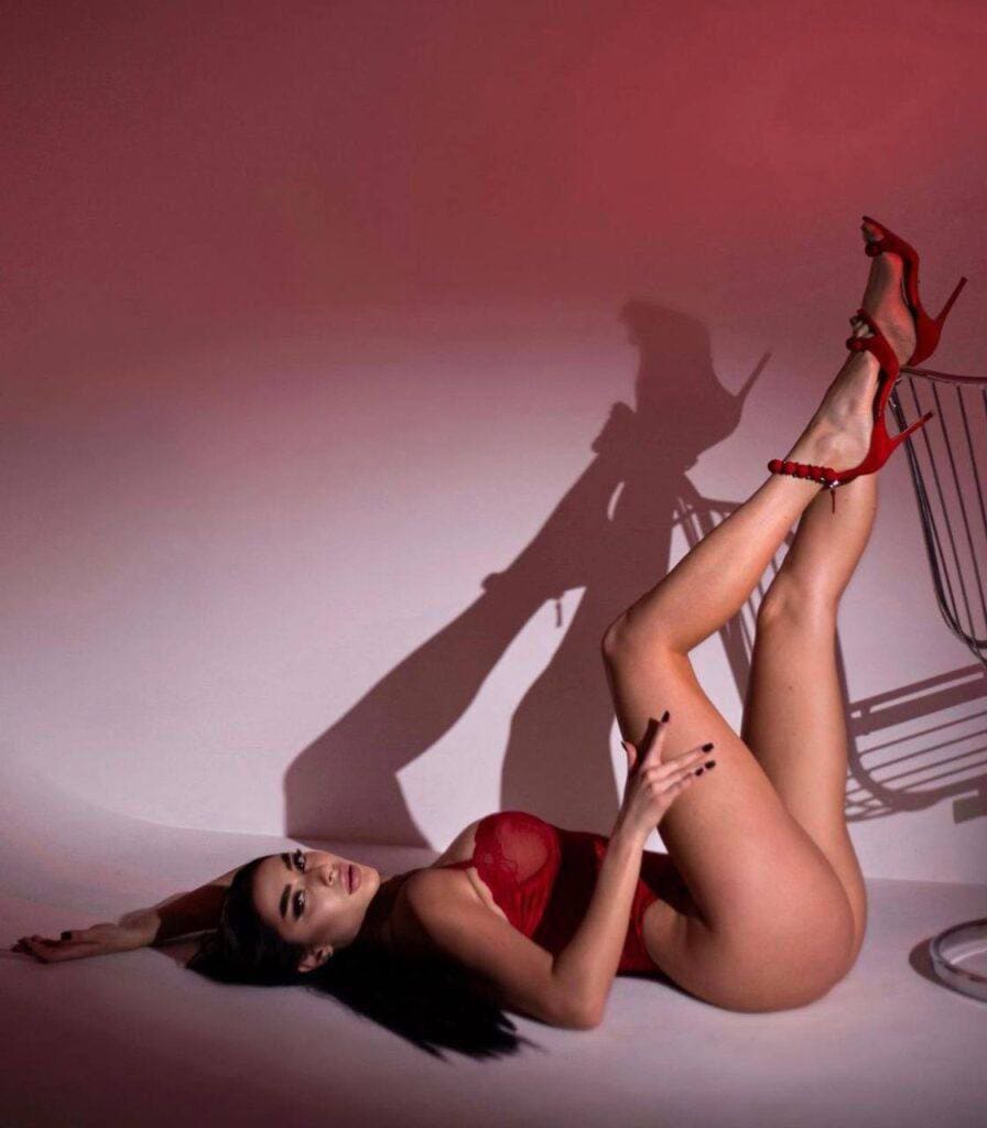 A woman lies on her side on a light floor in a red dress and heels, legs crossed against a pink-lit wall casting a shadow.