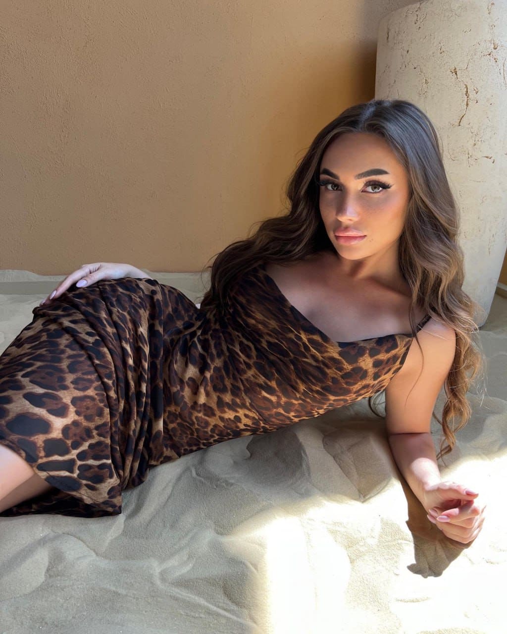 Woman lying on sand in a leopard-print dress, propped on one arm, looking at the camera near a stone pillar and tan wall.