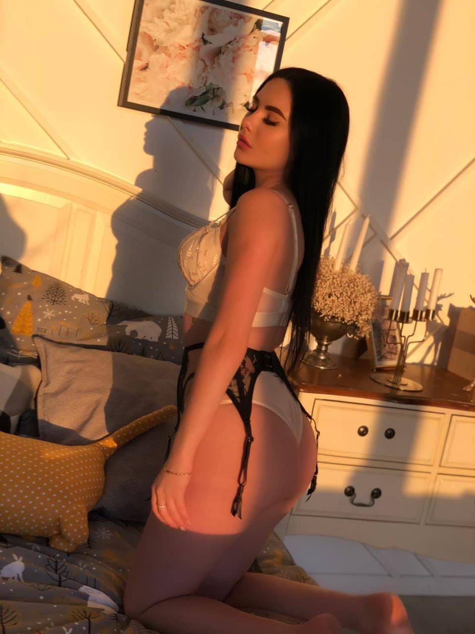 A woman kneels on a bed in warm golden light, wearing a white lace bra, high-cut underwear, and black lace garter straps, head turned to the side.