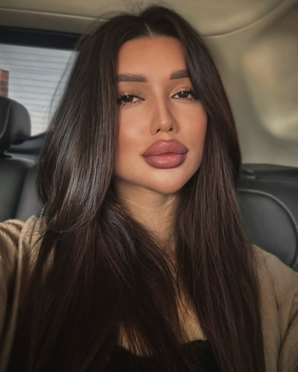 A close-up portrait of a woman with straight dark hair seated in a car, wearing a tan cardigan over a black top.