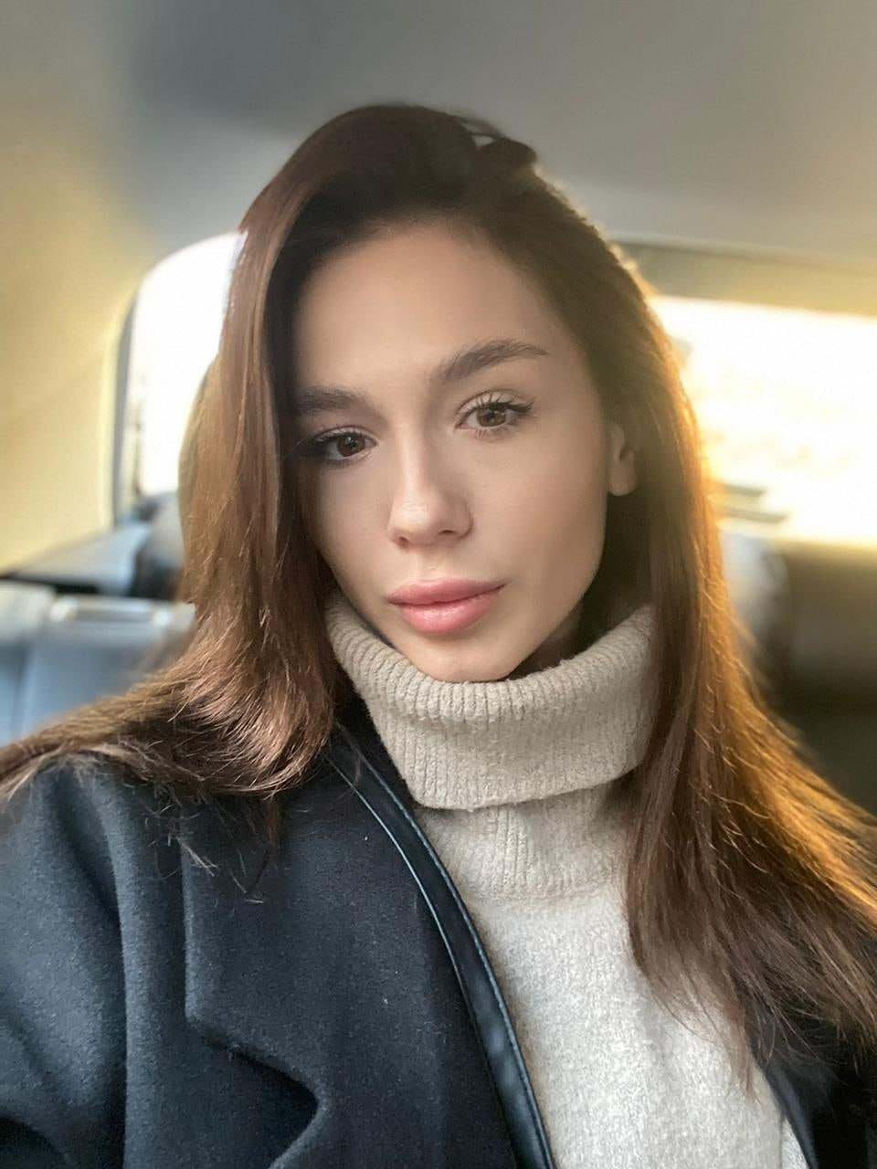 Seated inside a car, a woman in a cream turtleneck and dark coat looks toward the camera.