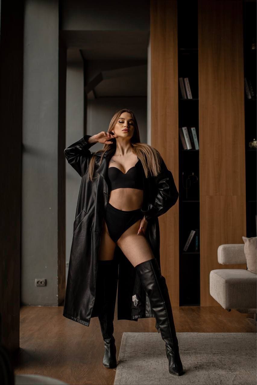 A woman with long brown hair stands in a room with wooden paneling, wearing an open black leather coat, bra, briefs, and thigh-high boots.