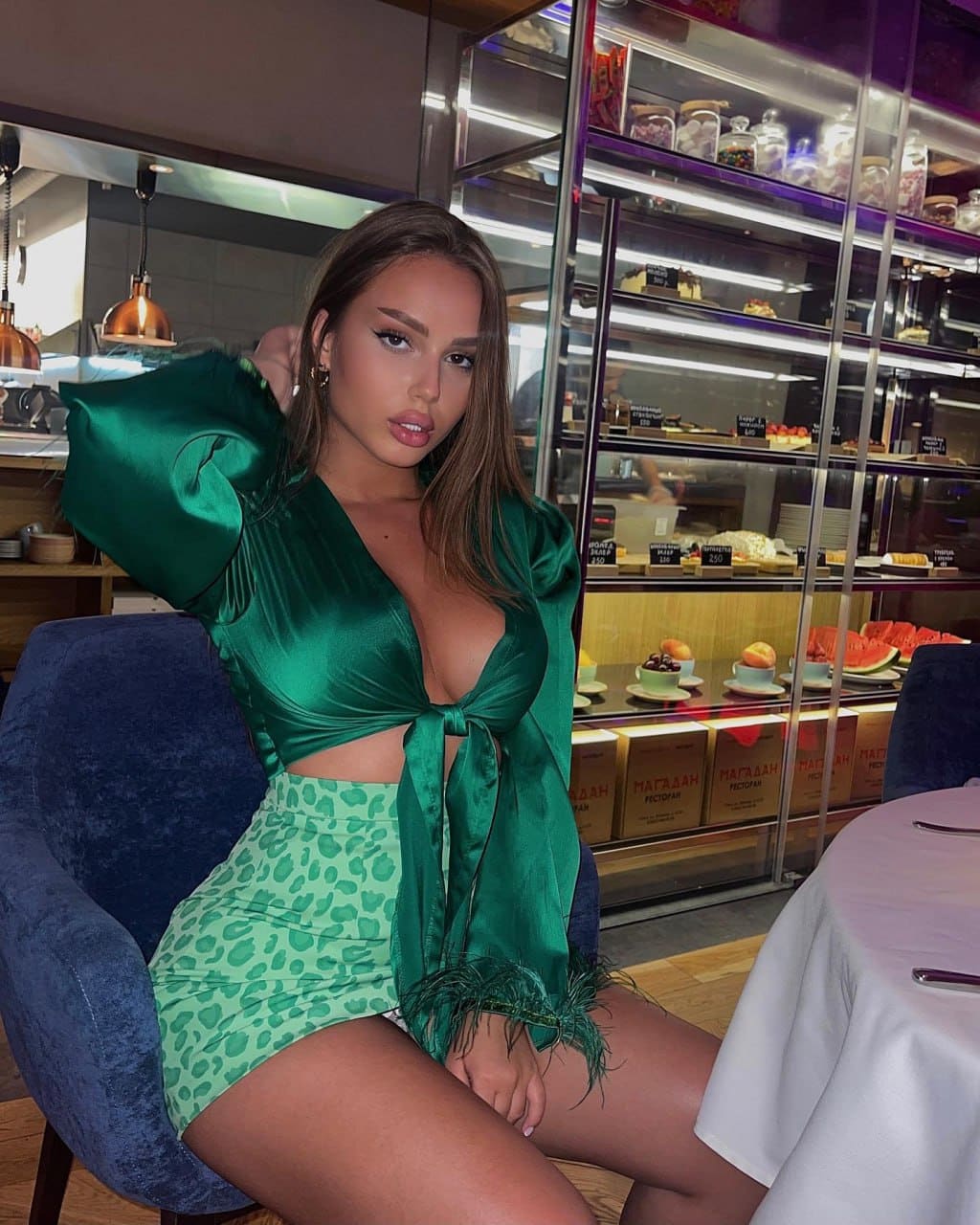 A woman sits on a blue velvet chair in a cafe, wearing a green satin top and leopard-print skirt, with pastries visible in a case behind her.