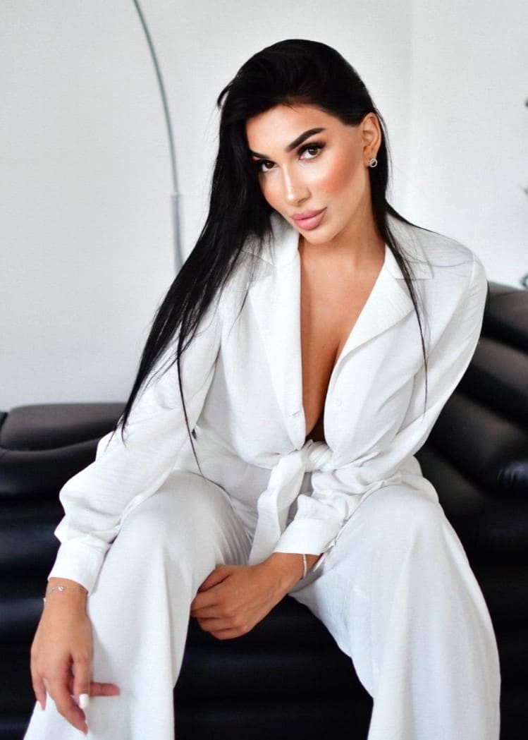 Naomi sits on a black leather couch wearing a white button-up shirt tied at the waist over white trousers in a bright room.