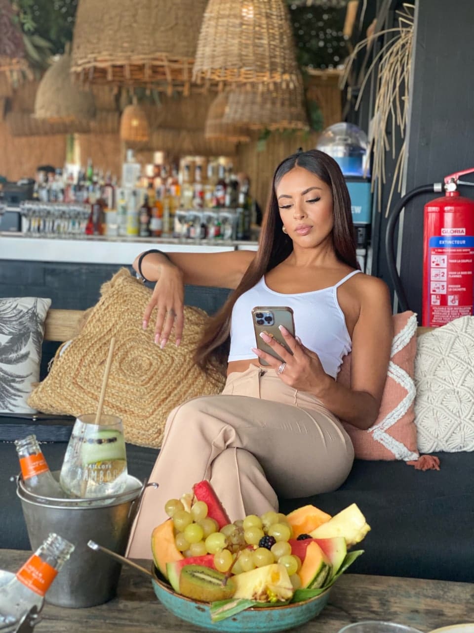 Lana sits on a couch in a white tank top and beige trousers, looking at her phone, with drinks and fruit on a table before her.