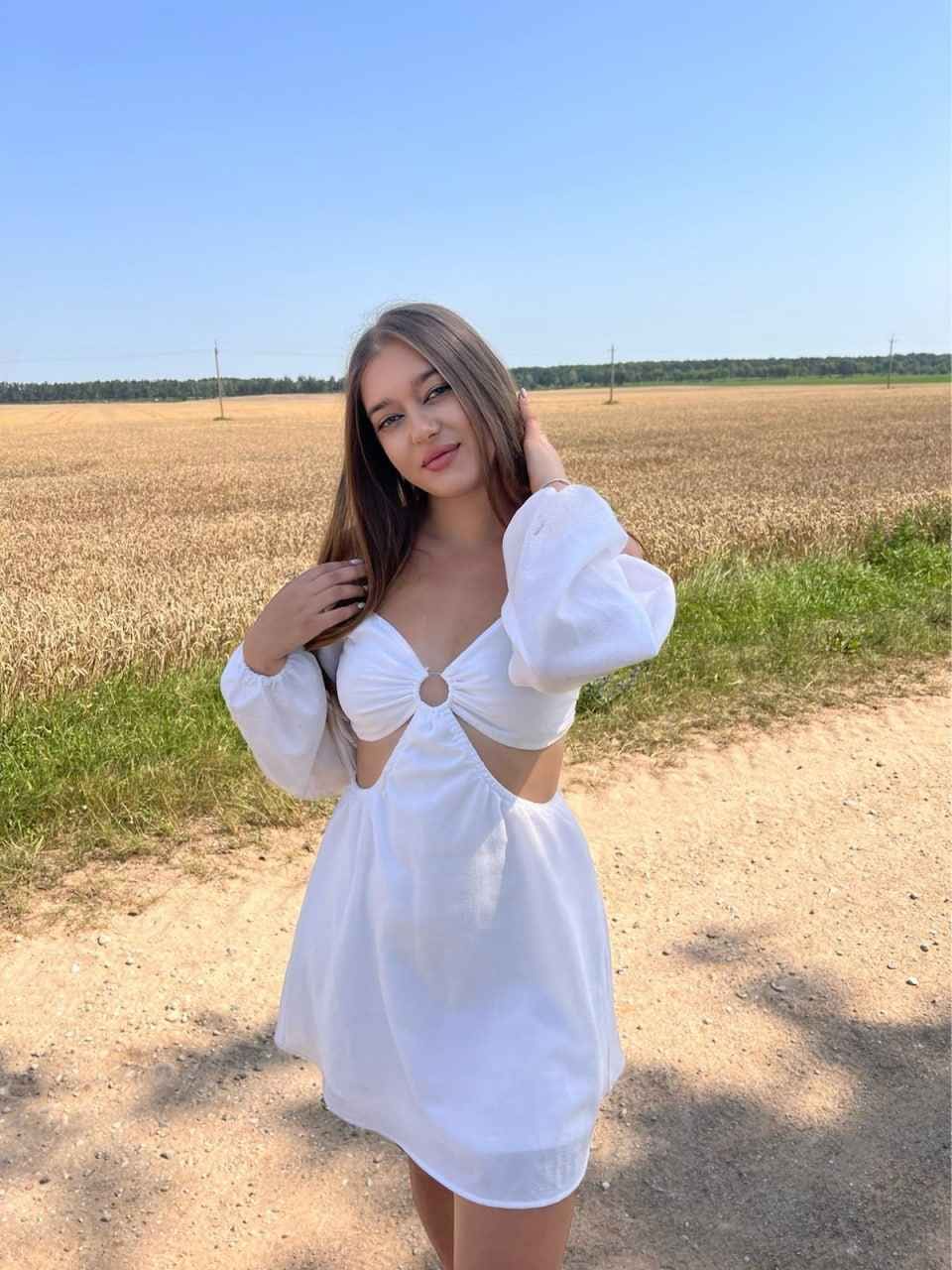 Chris stands on a dirt path by a harvested field in a white cut-out mini dress with puffed sleeves, one hand raised to her long brown hair.
