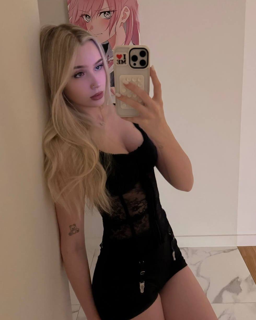 A blonde woman leans against a wall near an anime poster, taking a mirror selfie in a black lace-trimmed bodysuit and holding a patterned phone.