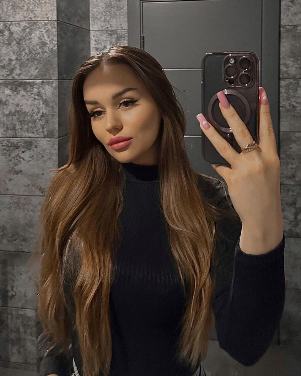 A woman takes a mirror selfie in a black high-neck top, holding a phone against a gray tiled wall.