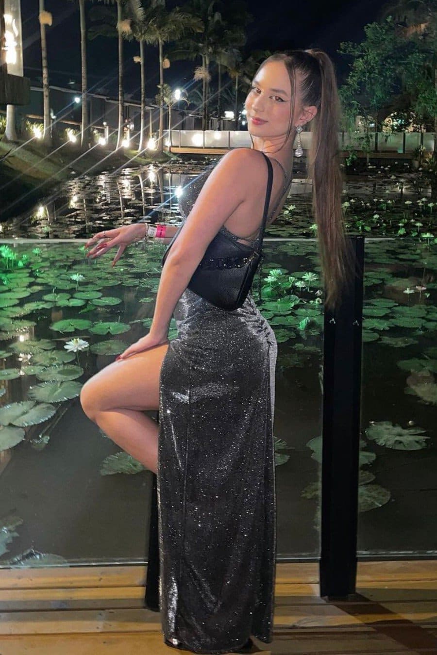 Woman in profile by a lily pond at night wears a black sequined dress with a high slit, one hand extended toward the water.