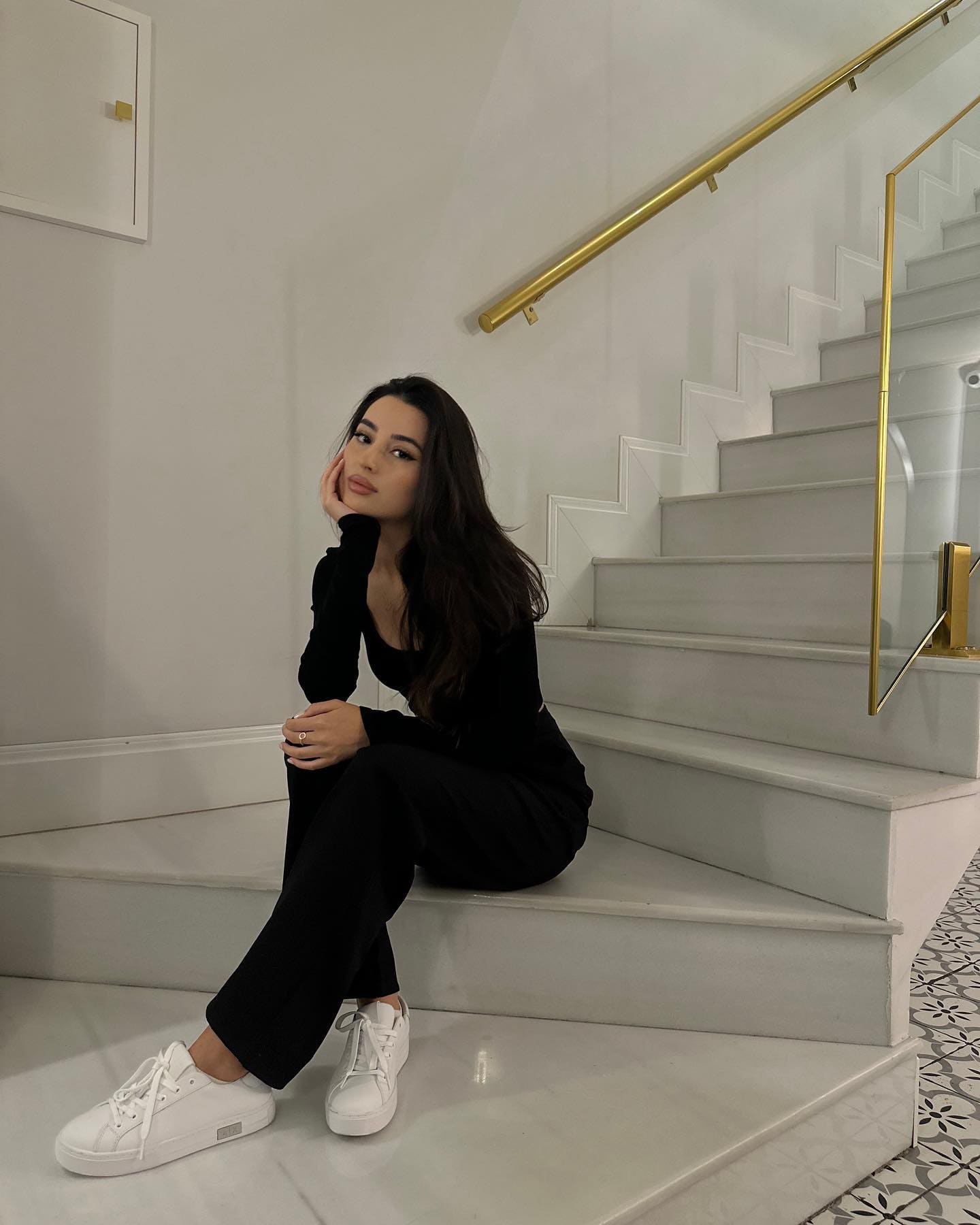 A woman sits on white marble stairs in a black top and trousers with white sneakers, hand on cheek, near a gold handrail and patterned floor.