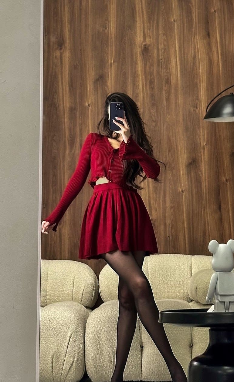 A woman in a red cropped cardigan, pleated mini skirt, and black tights stands by a cream sofa, holding a phone over her face.