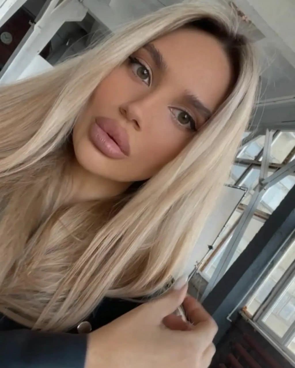 Close-up selfie of a platinum-blonde person in a dark top holding a small object, with windows and beams in the background.